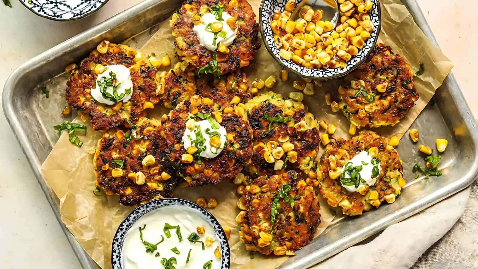 Corn fritters topped with sour cream and herbs, arranged on a parchment-lined tray with bowls of roasted corn and sour cream.