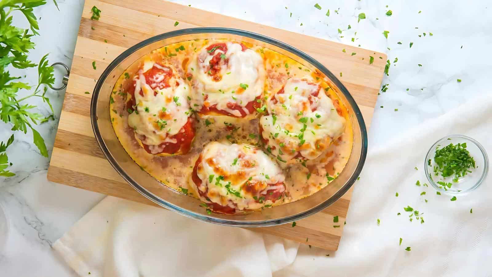 Clear glass dish with four cheese-topped stuffed peppers on a wooden board, sprinkled with chopped parsley, beside a small bowl of extra parsley garnish.