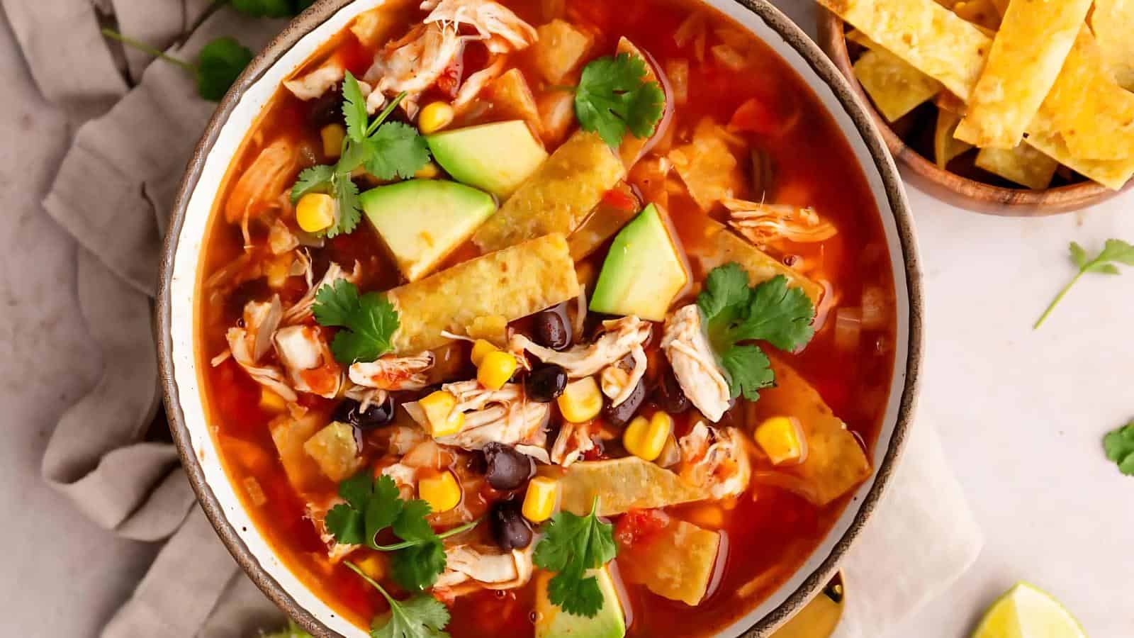 A bowl of tortilla soup with shredded chicken, avocado chunks, tortilla strips, black beans, corn, and cilantro. A small bowl of tortilla strips is on the side.