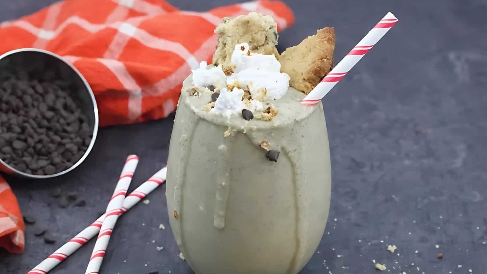 A creamy milkshake topped with whipped cream, cookie pieces, and chocolate chips in a glass with a red-striped straw. An orange cloth and spilled chocolate chips are in the background.