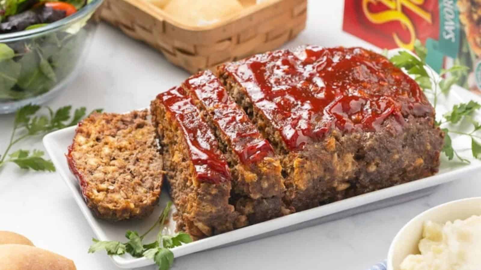 A glazed meatloaf partially sliced on a white rectangular plate, garnished with parsley, surrounded by salad, rolls, and boxed stuffing.