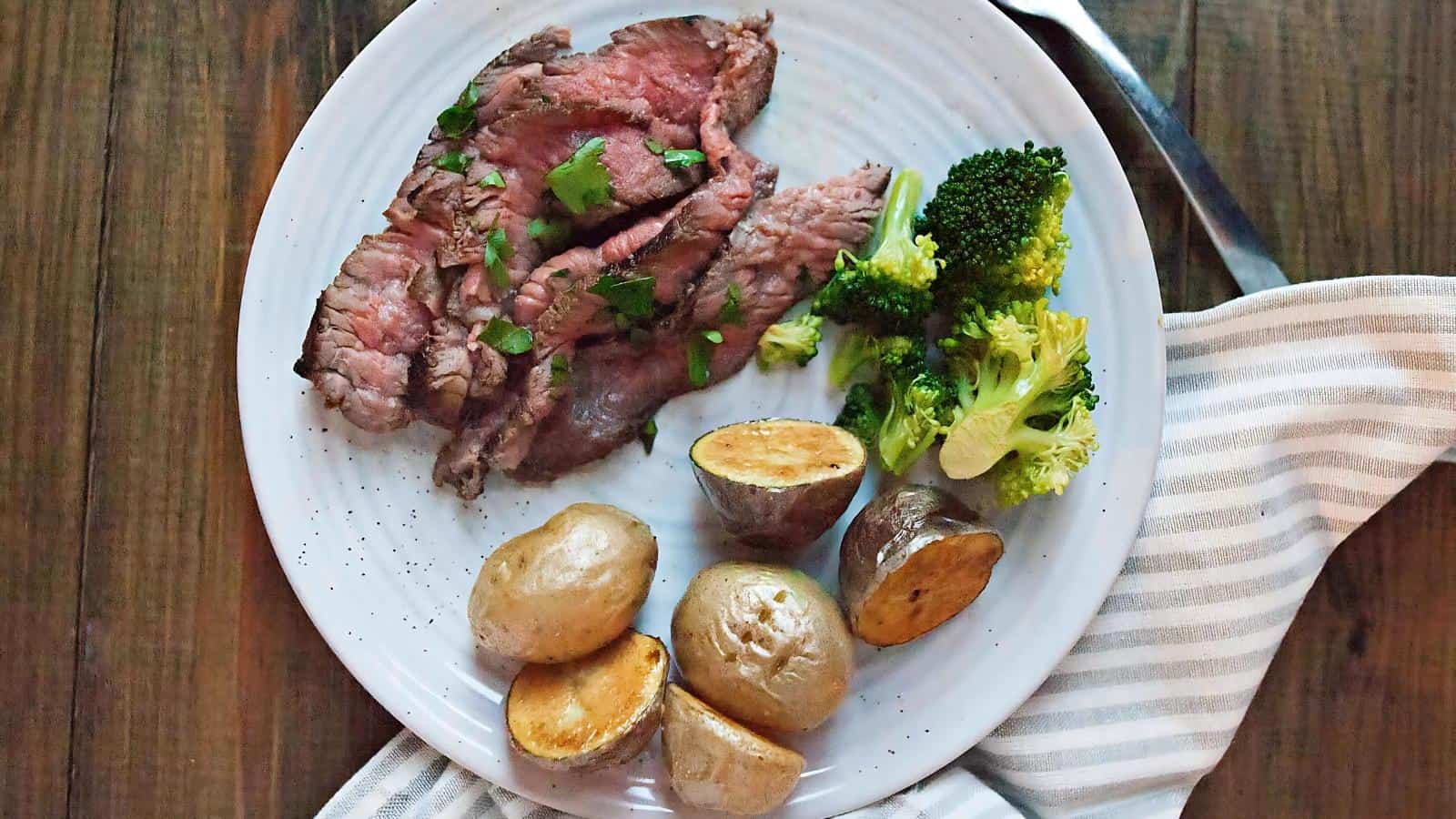 Plate with sliced steak, roasted potatoes, and steamed broccoli on a white plate, set on a wooden table with a striped napkin.