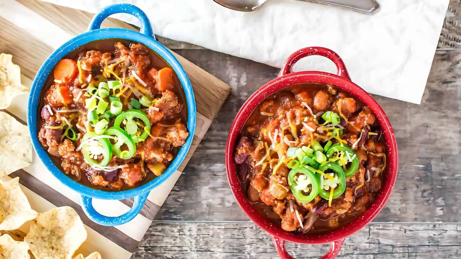 Two bowls of chili, one blue and one red, garnished with sliced green peppers, cheese, and scallions. Tortilla chips and a spoon are placed nearby on a wooden surface.