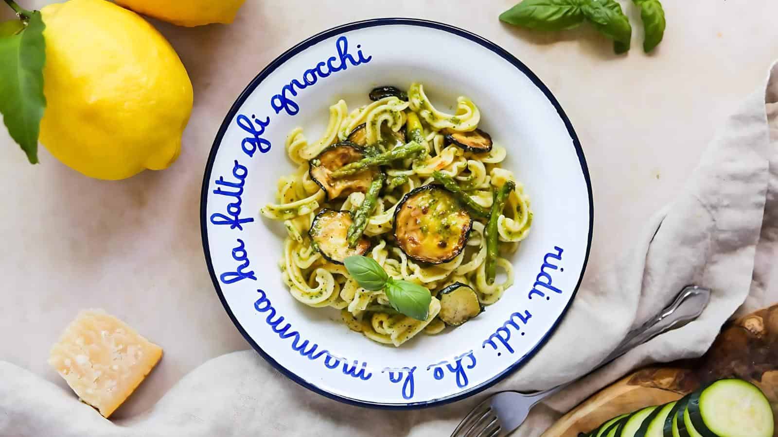 A plate of spiral pasta with zucchini and creamy sauce, garnished with basil. Lemons and parmesan are nearby, along with a fork on a cloth napkin. The plate has Italian text on the rim.