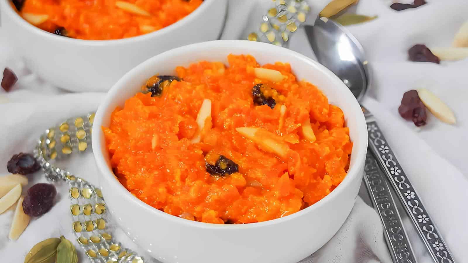 A white bowl filled with vibrant orange carrot halwa topped with raisins and slivered almonds, surrounded by scattered nuts and a spoon on a light background.