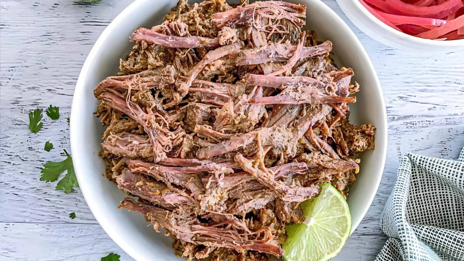 A bowl of shredded beef with a slice of lime on the side, placed on a white wooden surface.