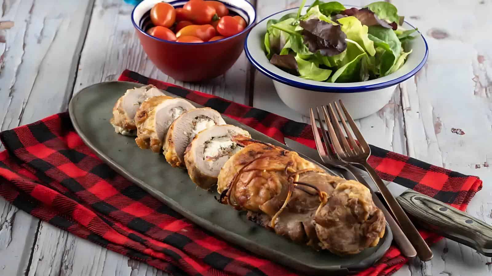 Sliced stuffed chicken breast on a black platter, placed on a red-and-black checkered cloth, with a fork and knife. Bowls of cherry tomatoes and mixed greens are in the background.