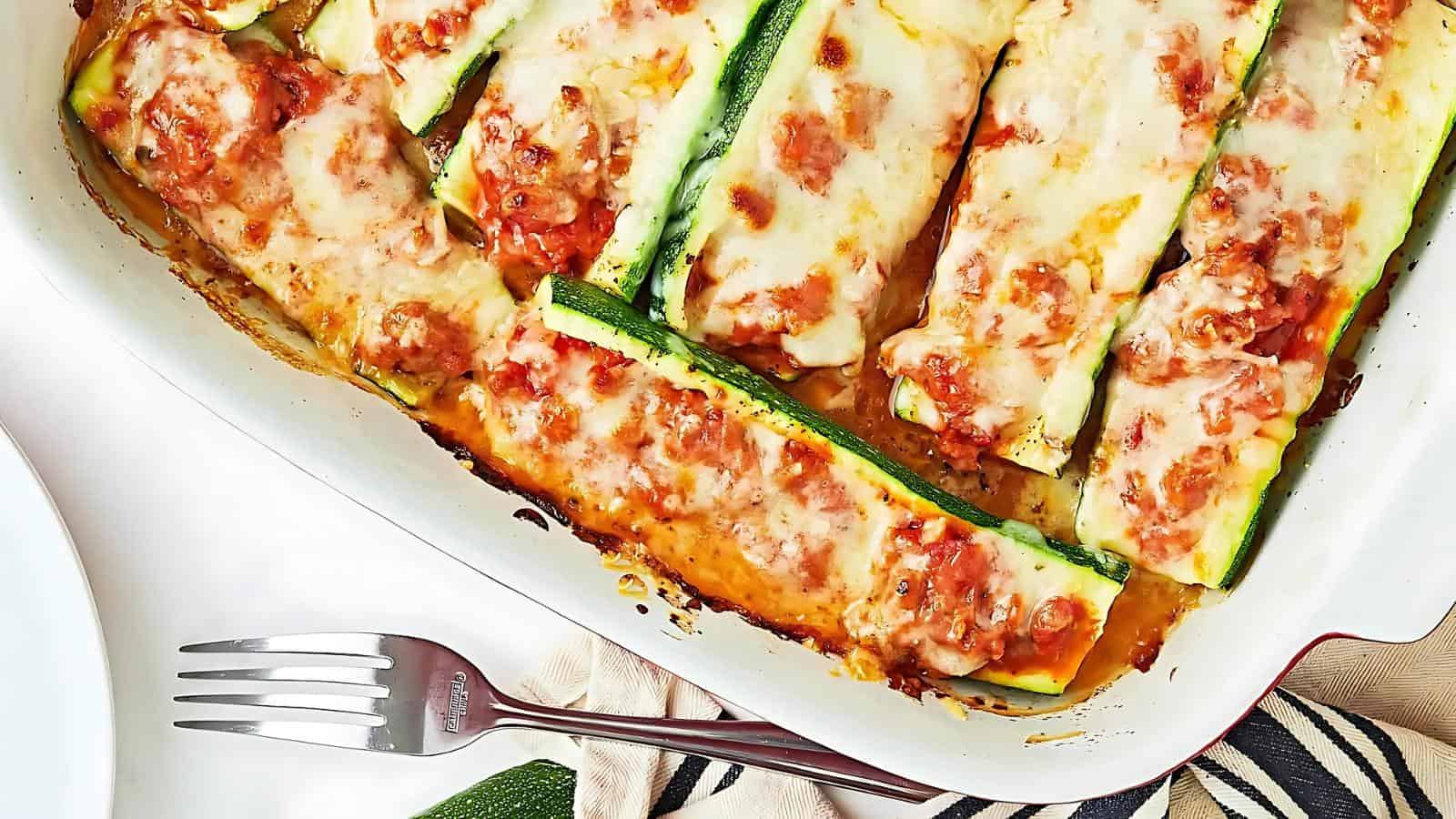 Baked zucchini boats filled with a tomato and cheese mixture in a white dish, next to a fork on a striped cloth.