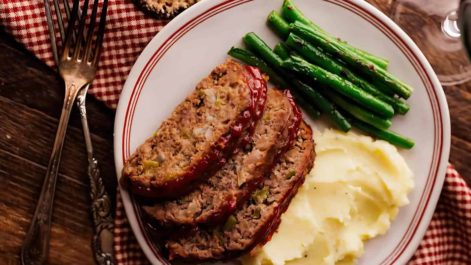 A plate with three slices of glazed meatloaf, mashed potatoes, and green beans on a red and white checkered cloth.