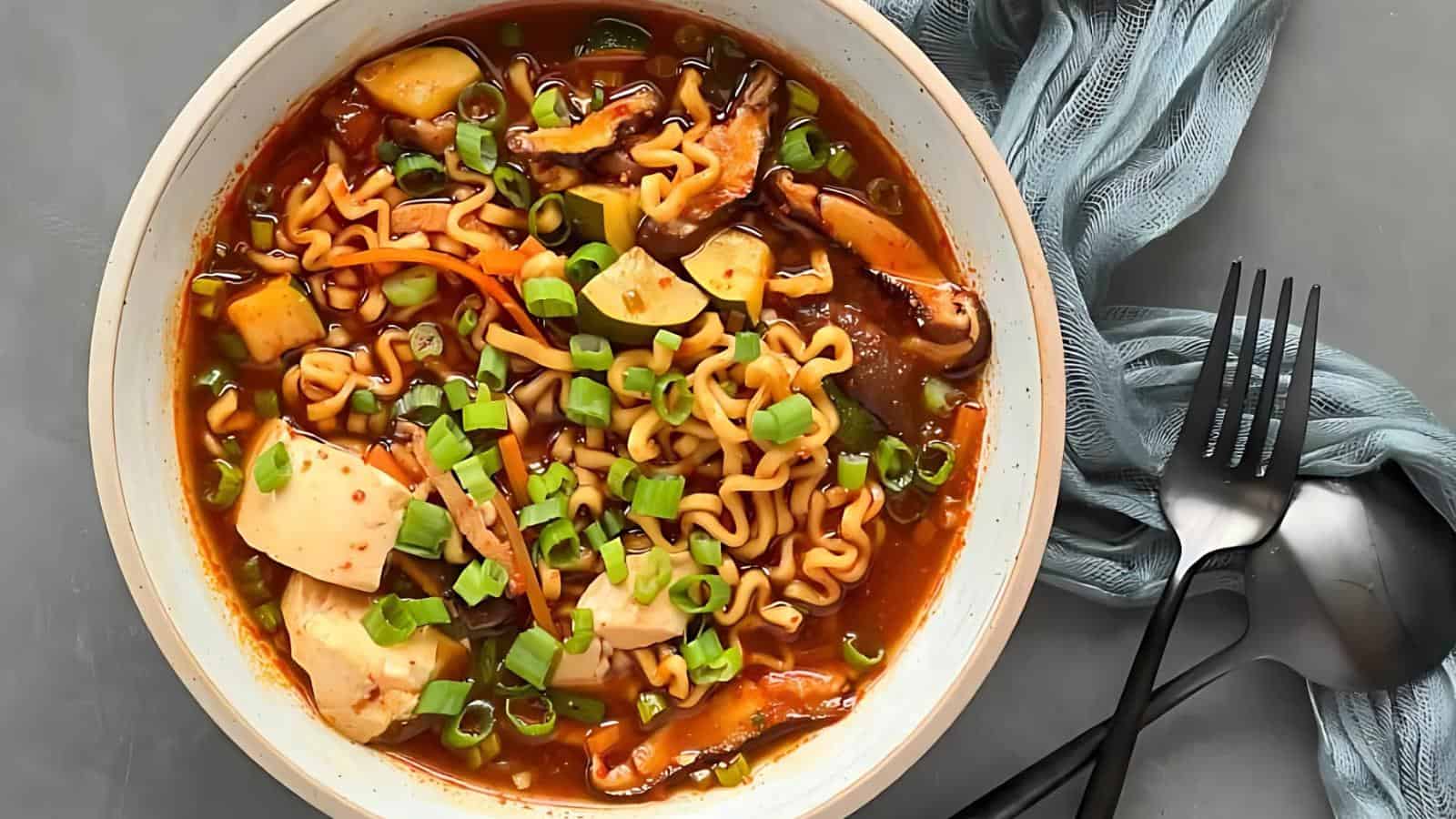 A bowl of spicy ramen topped with tofu, mushrooms, and chopped green onions. A blue cloth and a fork are placed beside the bowl.