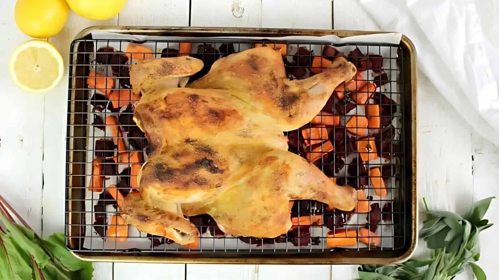 Roasted spatchcock chicken on a wire rack over diced carrots and onions on a baking sheet. Lemons and greens are visible on the side.