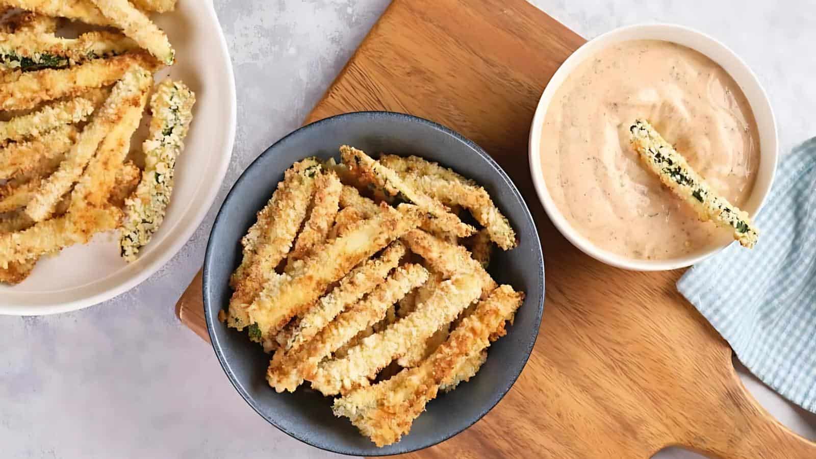 Crispy breaded zucchini fries in a black bowl, with a separate white bowl of dipping sauce on a wooden board.