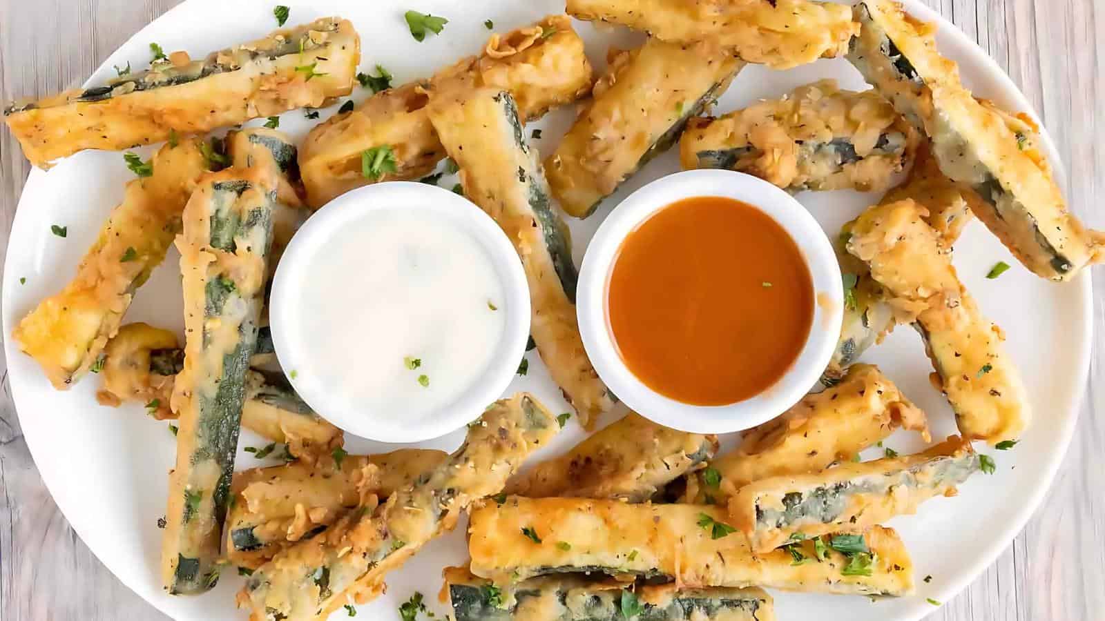 A plate of tempura-fried zucchini sticks is served with two dipping sauces: one is creamy white, the other is orange.