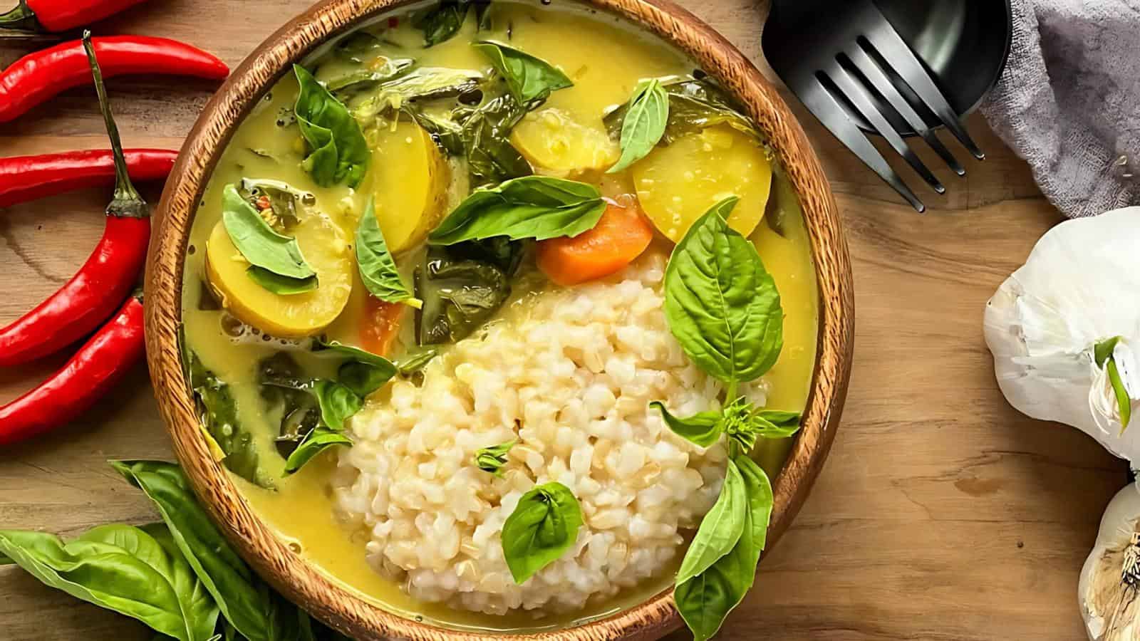 A bowl of creamy vegetable curry with rice, garnished with basil leaves. Red chilies and garlic are placed around the wooden table setting.