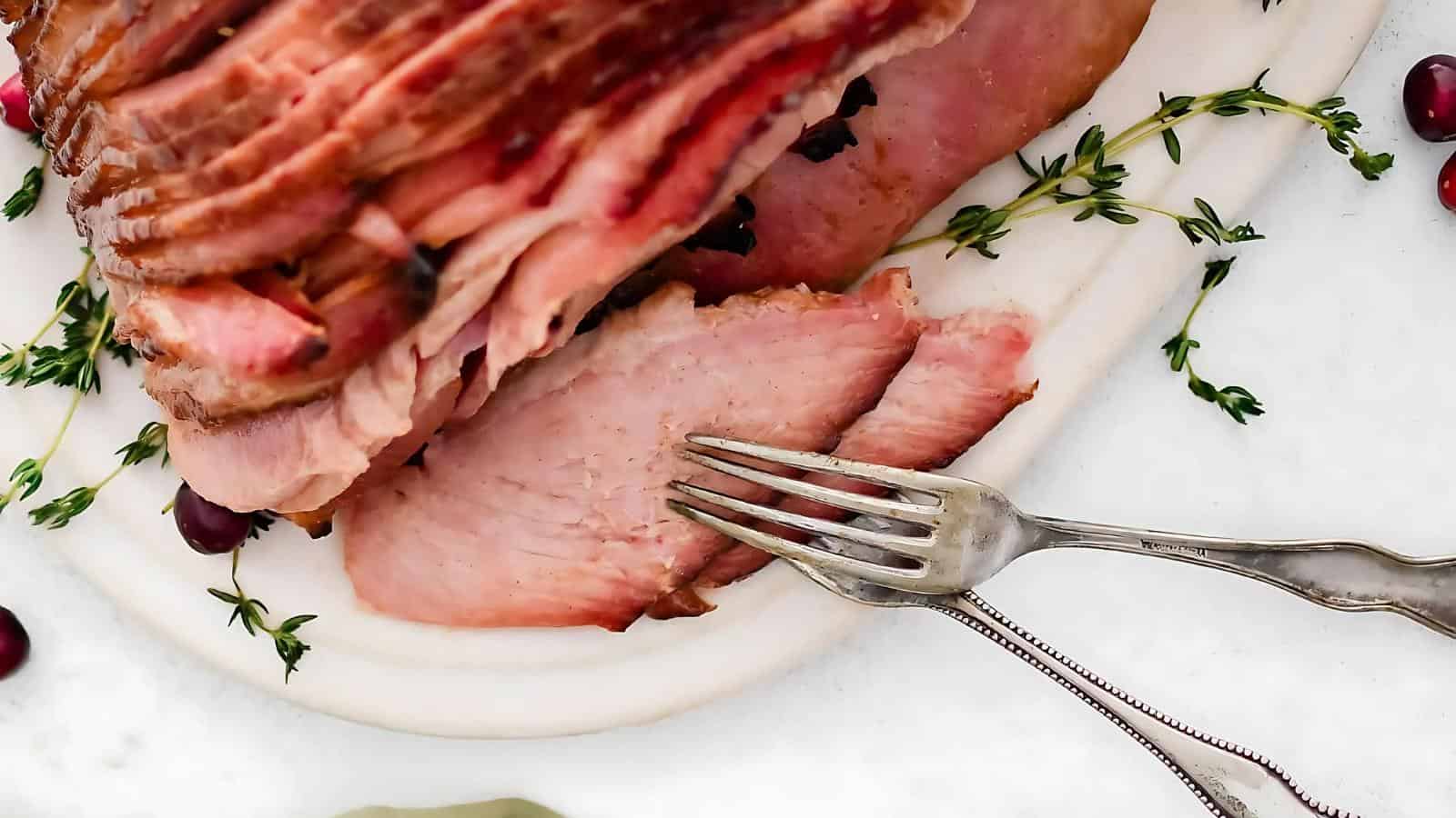 Sliced ham on a white plate with a fork beside it, garnished with fresh thyme and cranberries.