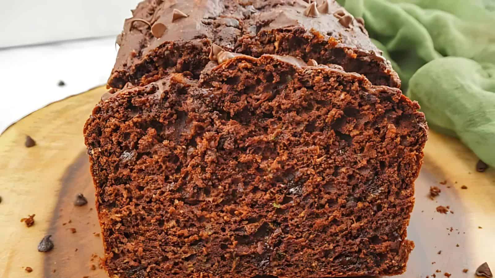 A close-up of a sliced chocolate loaf cake on a wooden board, with chocolate chips visible on top and a green cloth in the background.