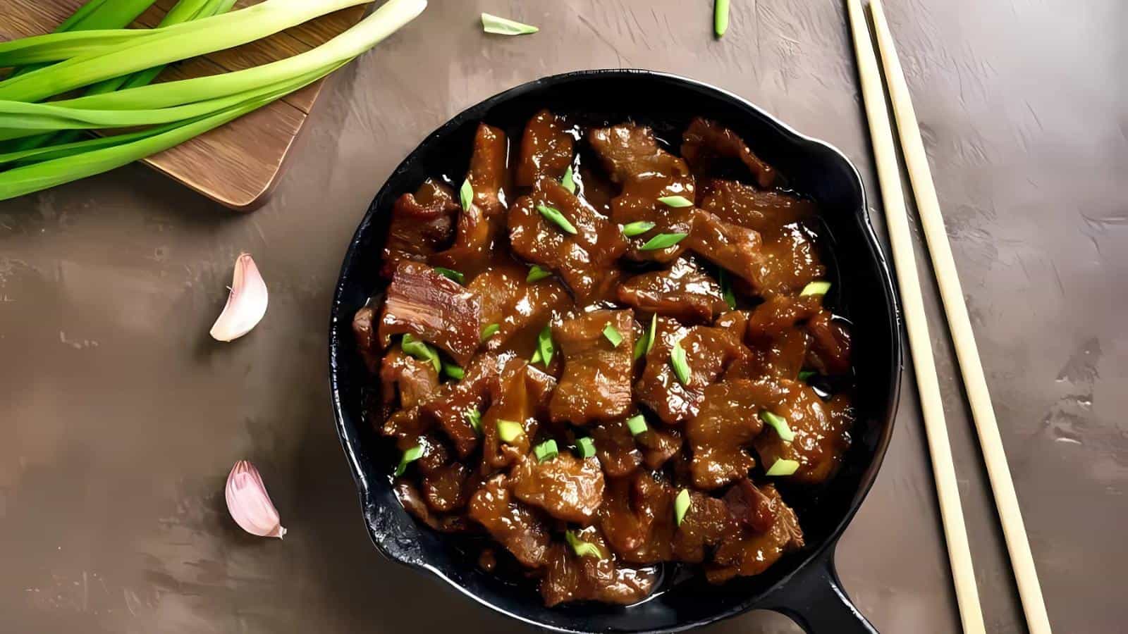 A black skillet with cooked beef slices coated in a glossy sauce, garnished with chopped green onions. Nearby are chopsticks, green onions, and garlic cloves on a brown surface.