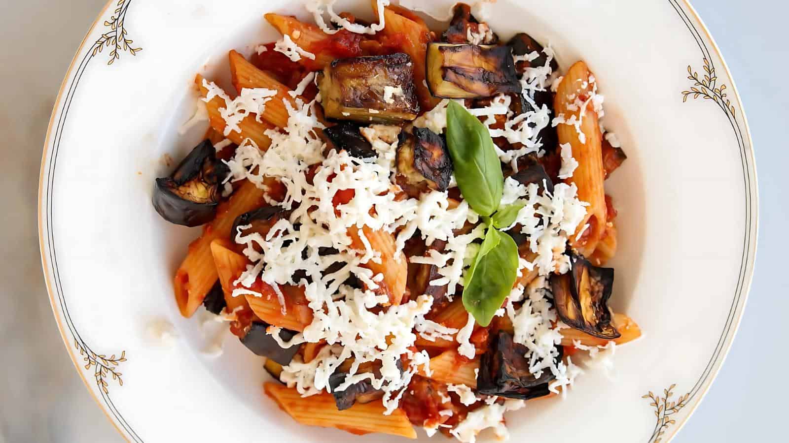 A plate of penne pasta with tomato sauce, topped with grilled eggplant, grated cheese, and basil leaves, served on a decorative white dish.