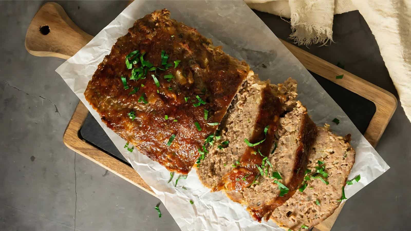 A sliced meatloaf garnished with chopped parsley on parchment paper, placed on a wooden cutting board.