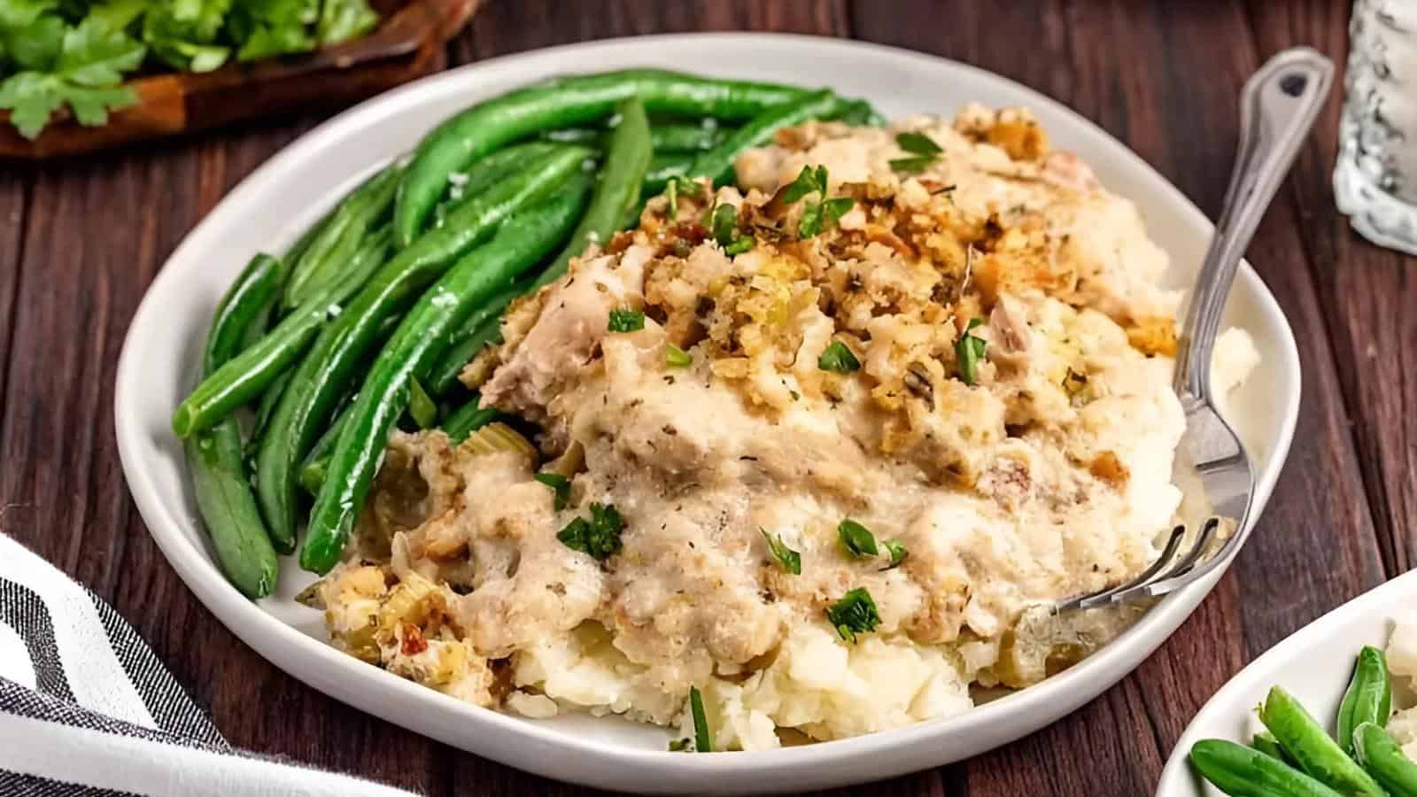 A plate of creamy chicken and stuffing over mashed potatoes, garnished with parsley, served with steamed green beans.