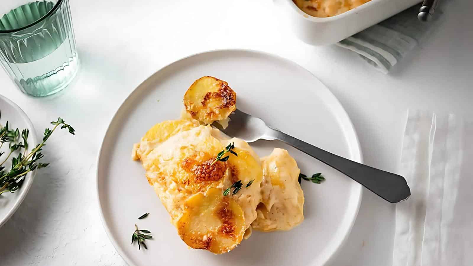A white plate with a serving of baked scalloped potatoes and a fork, garnished with fresh thyme. A glass of water and a dish of potatoes are in the background.