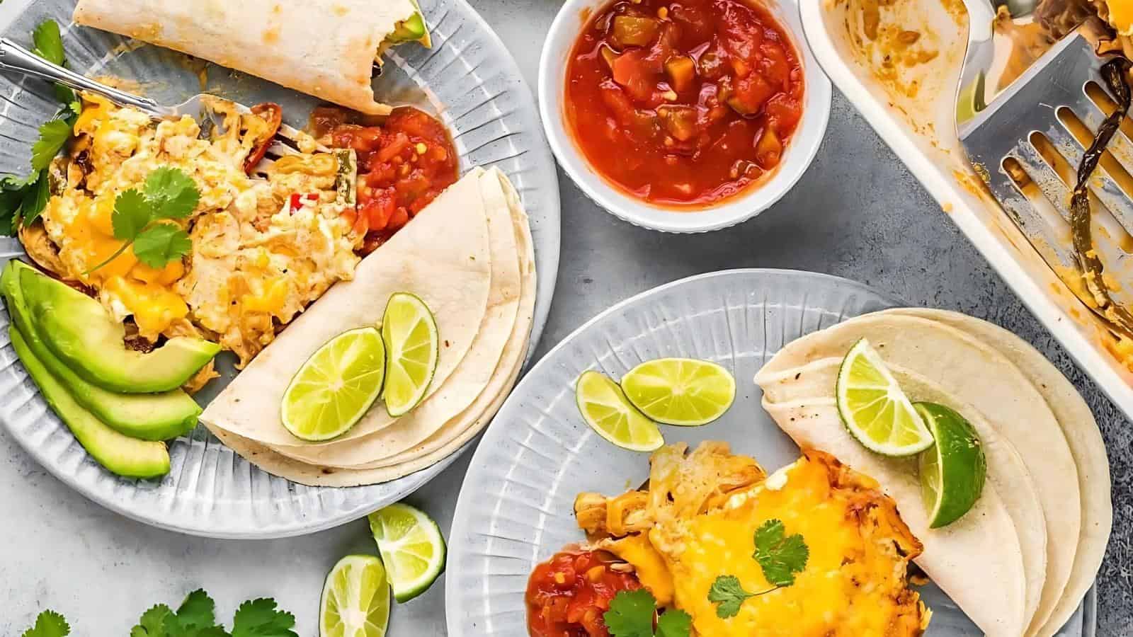 Two plates of enchiladas with melted cheese, avocado slices, lime wedges, and salsa on a gray surface. A side dish of salsa and a casserole dish with cooked enchiladas are visible.