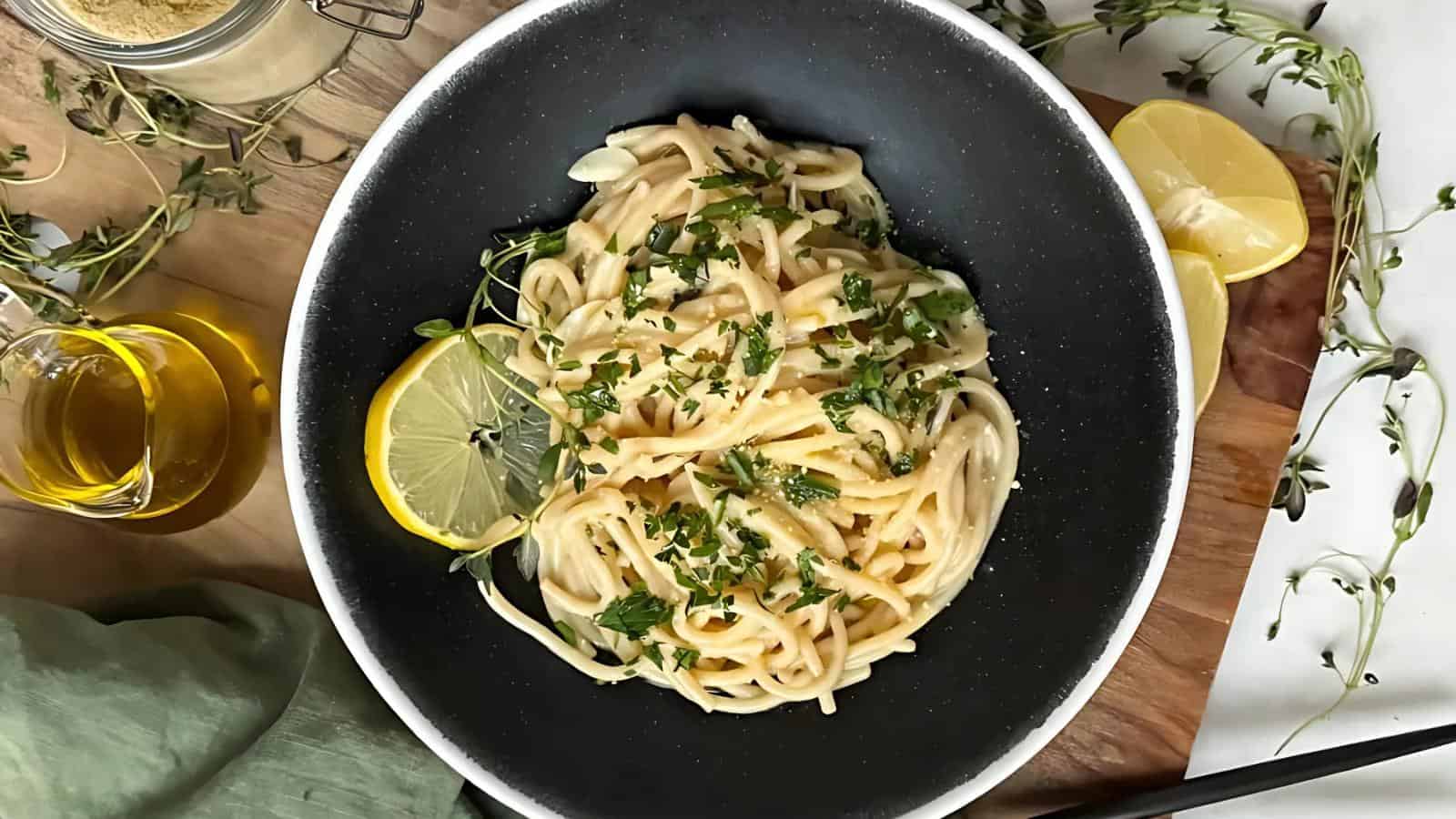 A black bowl of spaghetti aglio e olio garnished with parsley and a lemon slice. Olive oil and thyme are nearby on a wooden surface.