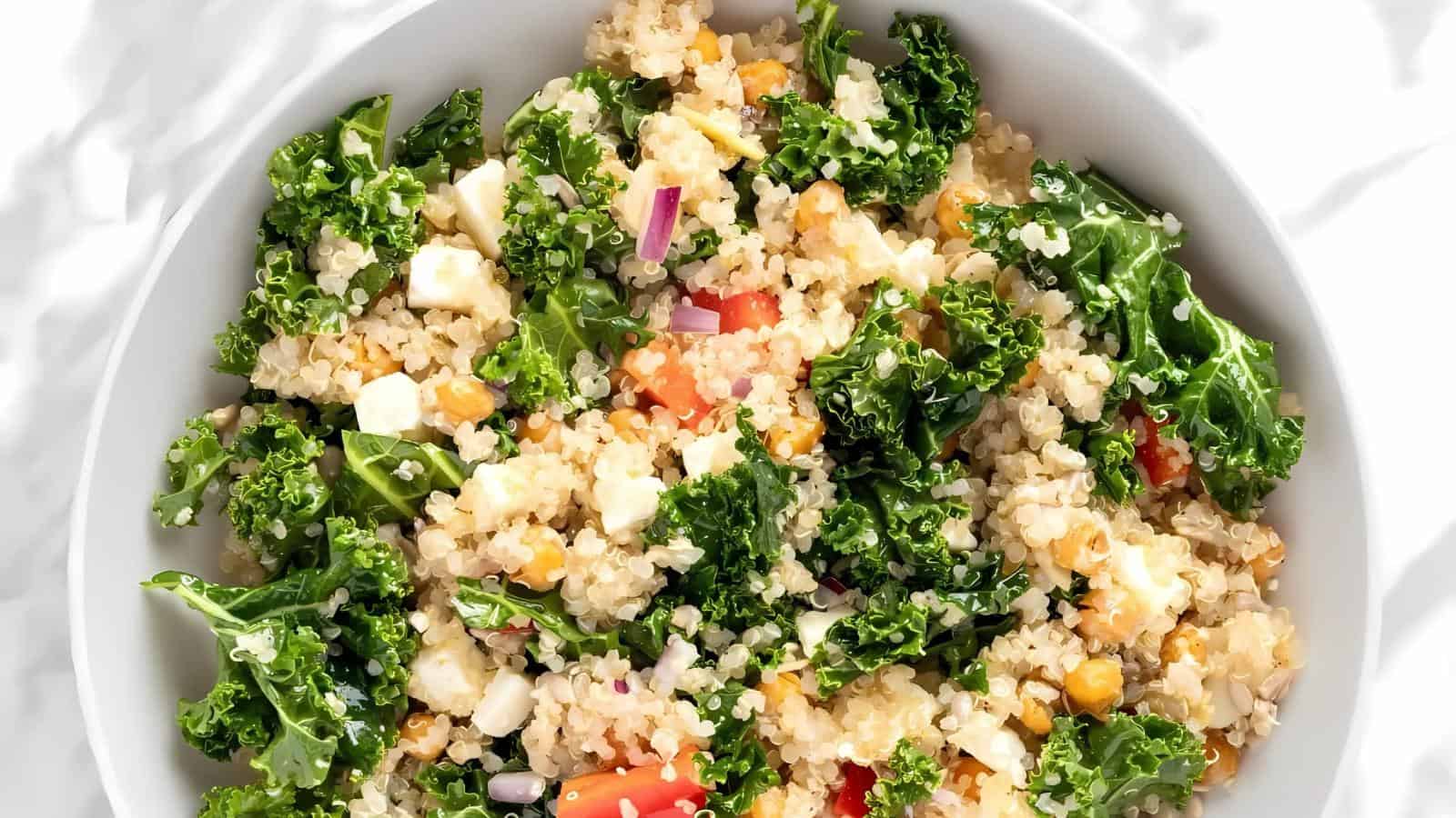 A white bowl filled with quinoa salad, mixed with kale, red onion, tomato, feta cheese, and chickpeas.