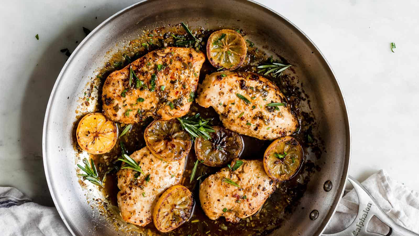 Pan with cooked chicken breasts, lemon slices, and herbs in a sauce.