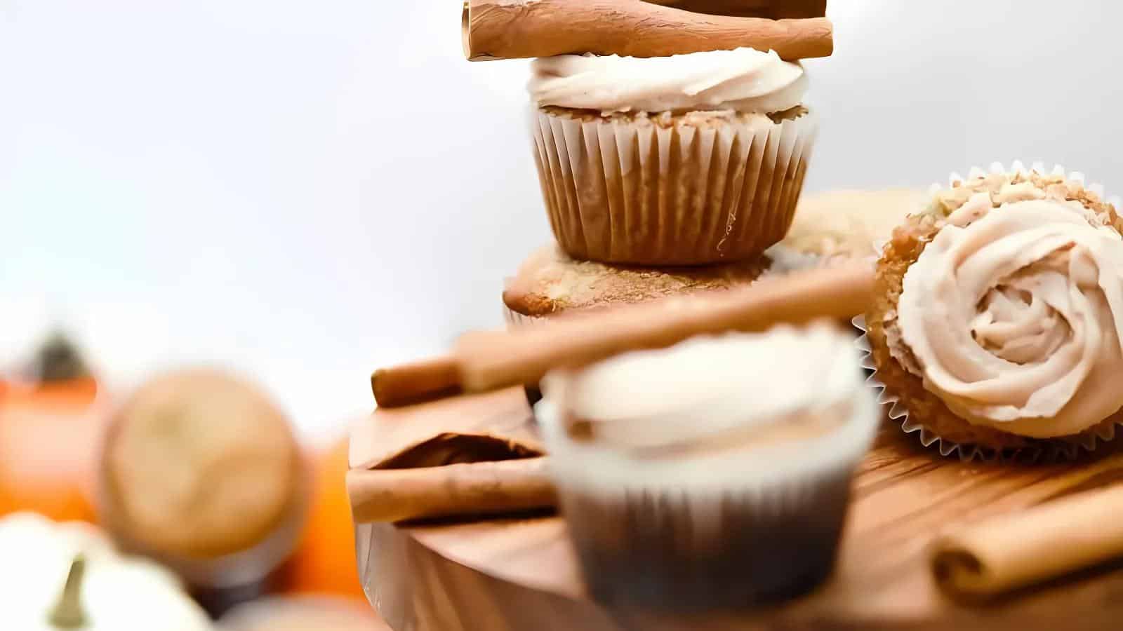 Cupcakes with white frosting and cinnamon sticks on a wooden board. Blurred background with additional cupcakes and small pumpkins.