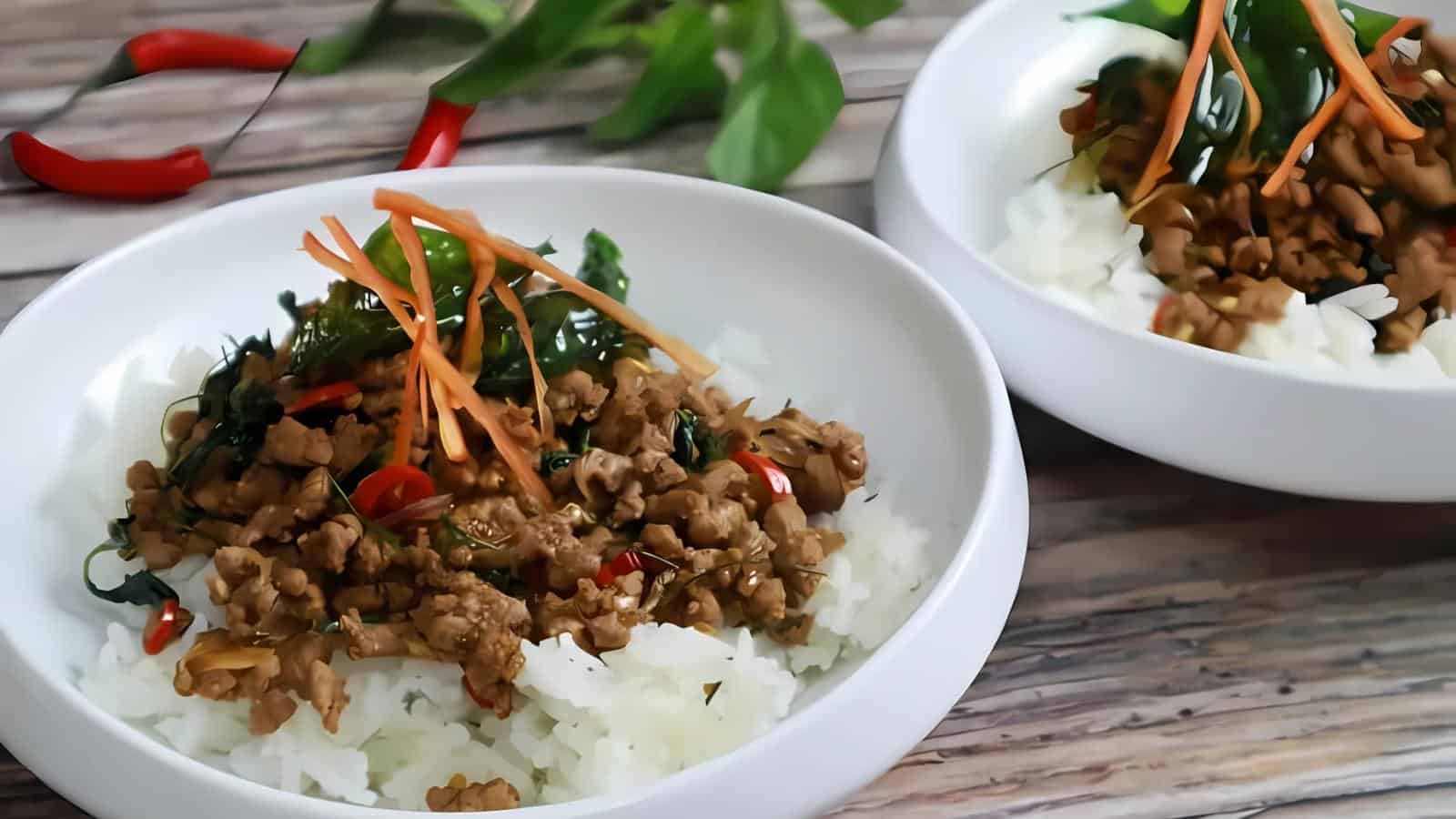 Two white bowls contain rice topped with stir-fried minced meat, vegetables, and garnished with shredded carrots and herbs. Red chili peppers are in the background on a wooden surface.