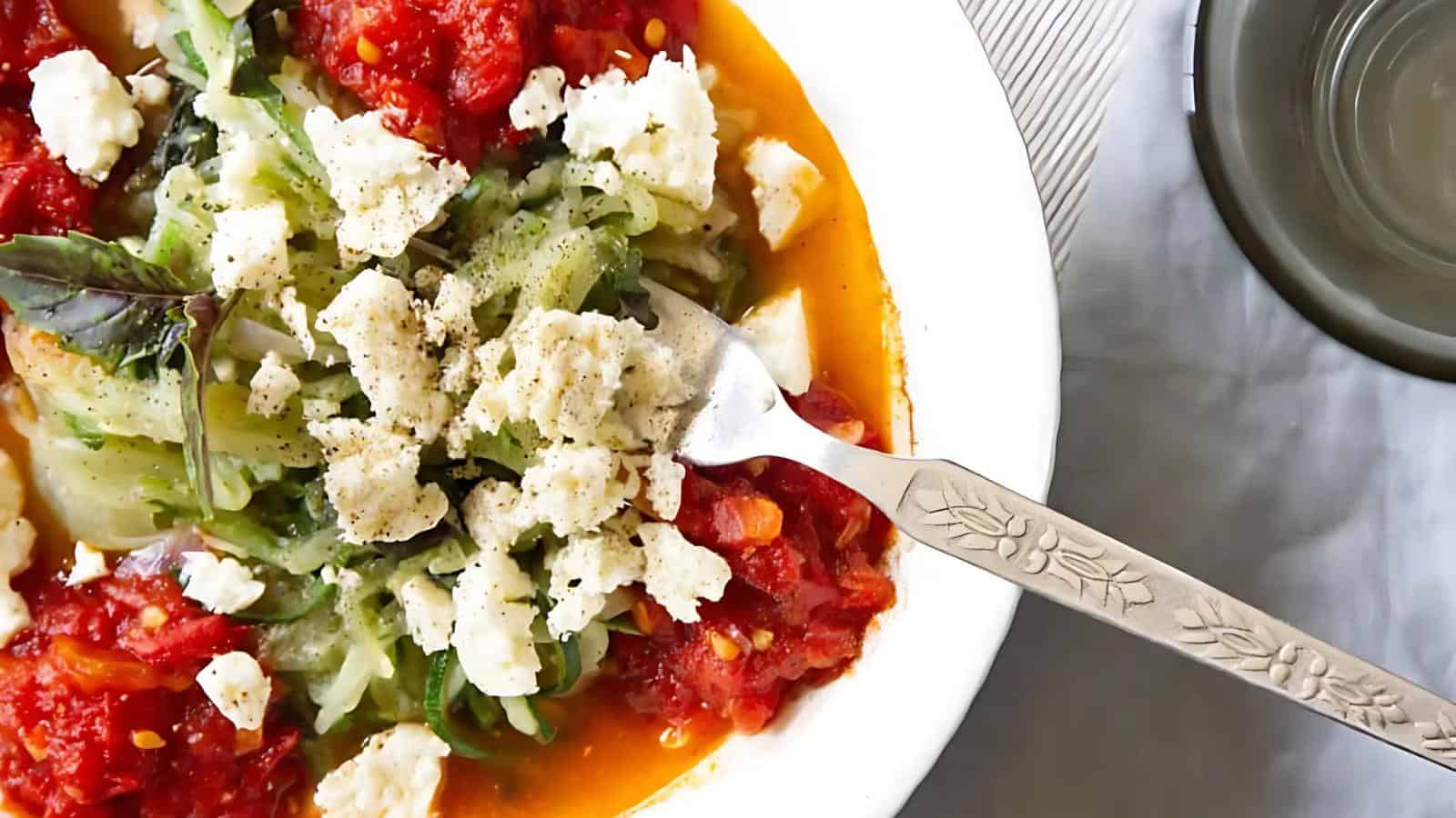 A bowl of zucchini noodles topped with tomato sauce, crumbled feta, and a basil leaf, with a fork on the side.