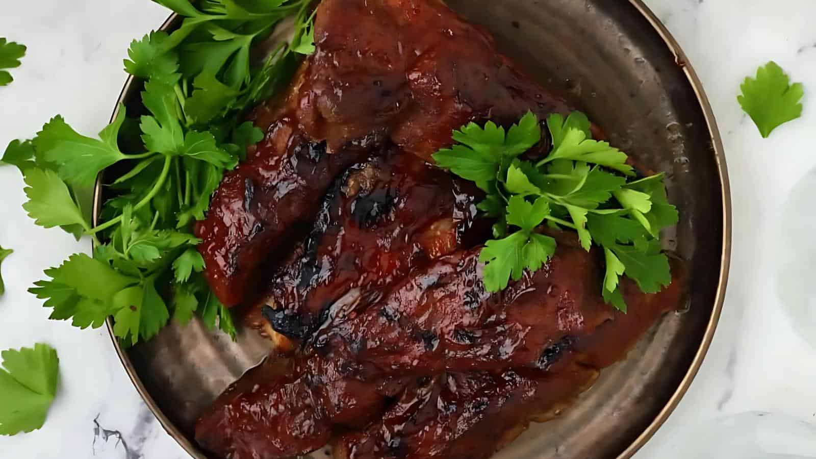 Barbecued ribs coated in sauce are served on a metal plate, garnished with fresh parsley leaves.