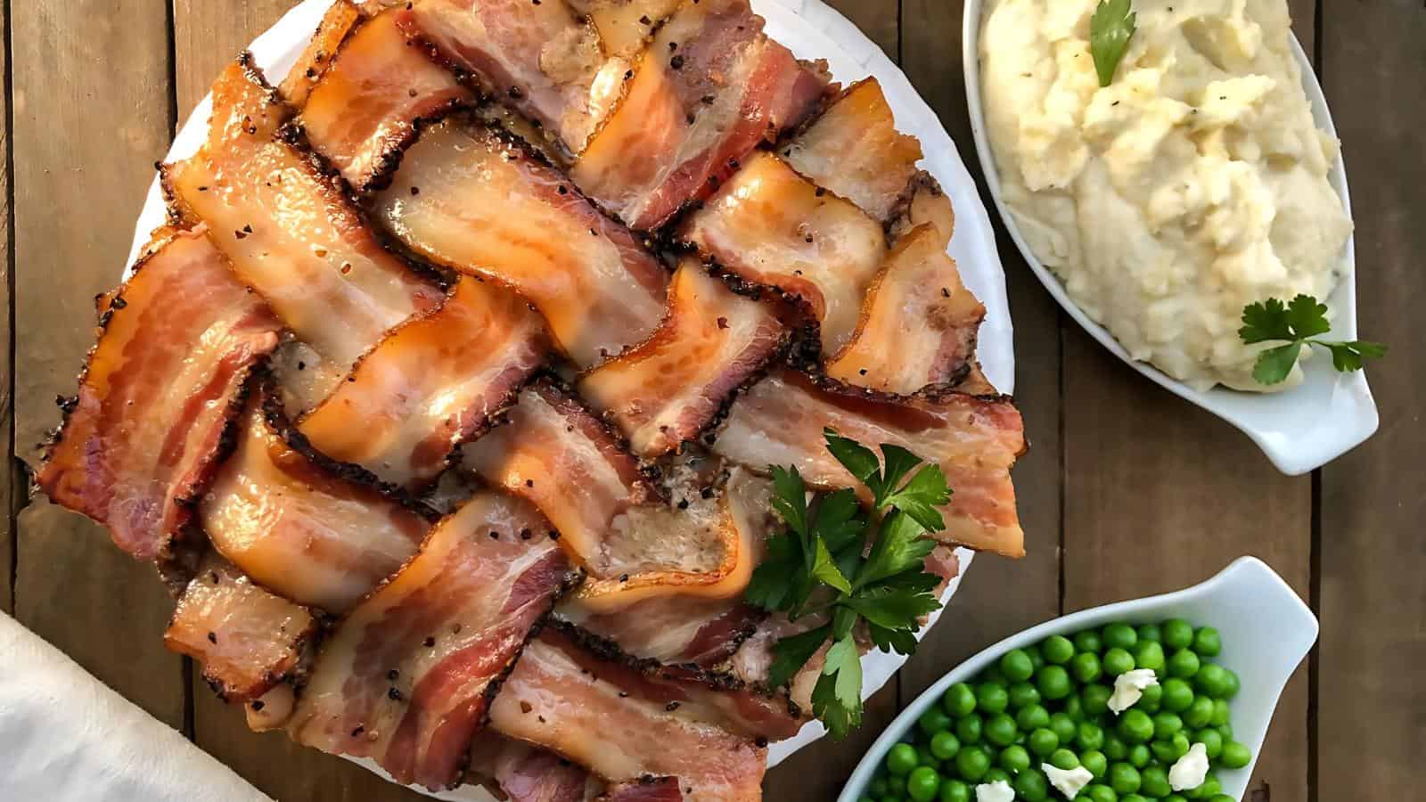 Bacon-wrapped dish with parsley garnish on a plate, alongside mashed potatoes and peas, on a wooden table.