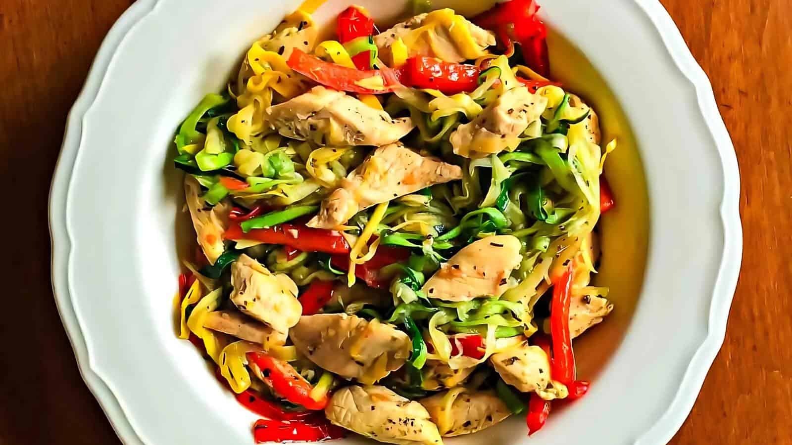 A white plate filled with zucchini noodles, sliced chicken, and red bell peppers, served on a wooden table.