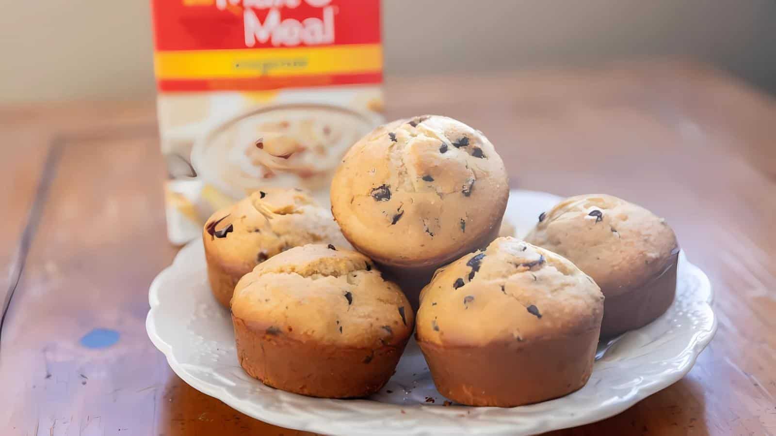 Five chocolate chip muffins on a white plate with a box of Cream of Wheat in the background.