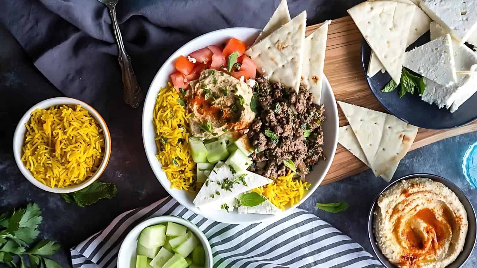 Bowl with yellow rice, beef, cucumber, tomato, tzatziki, and pita triangles. Surroundings include more rice, pita, feta, tzatziki, and parsley on a dark surface with striped cloth.