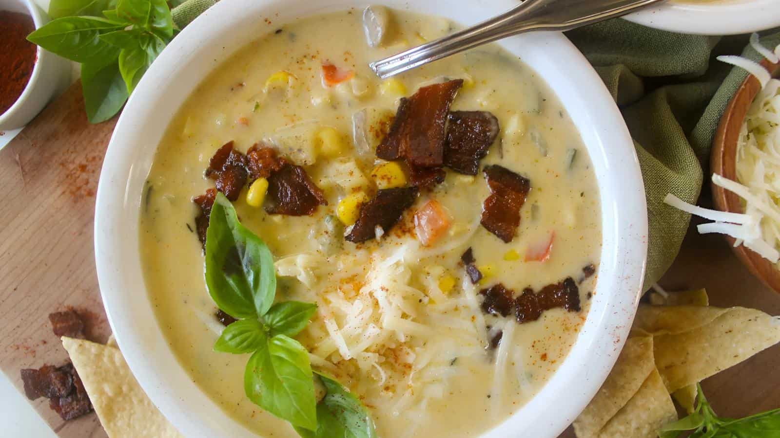 A bowl of creamy soup topped with bacon, cheese, and basil, with tortilla chips on the side.