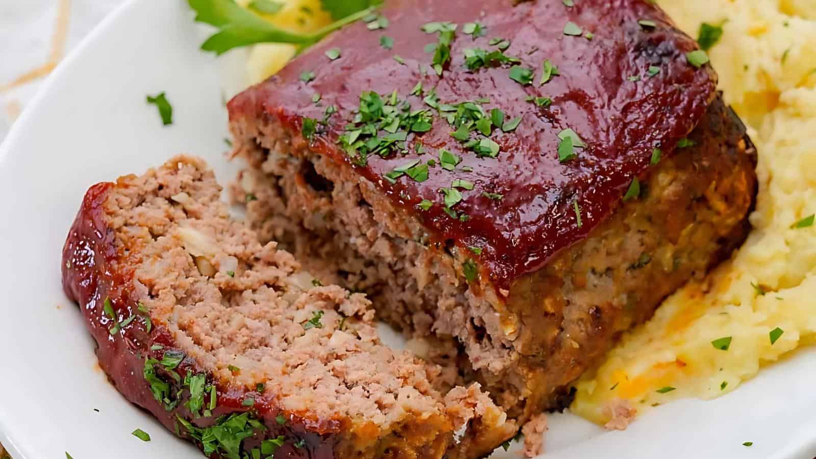 Slice of glazed meatloaf garnished with herbs, served with mashed potatoes on a white plate.