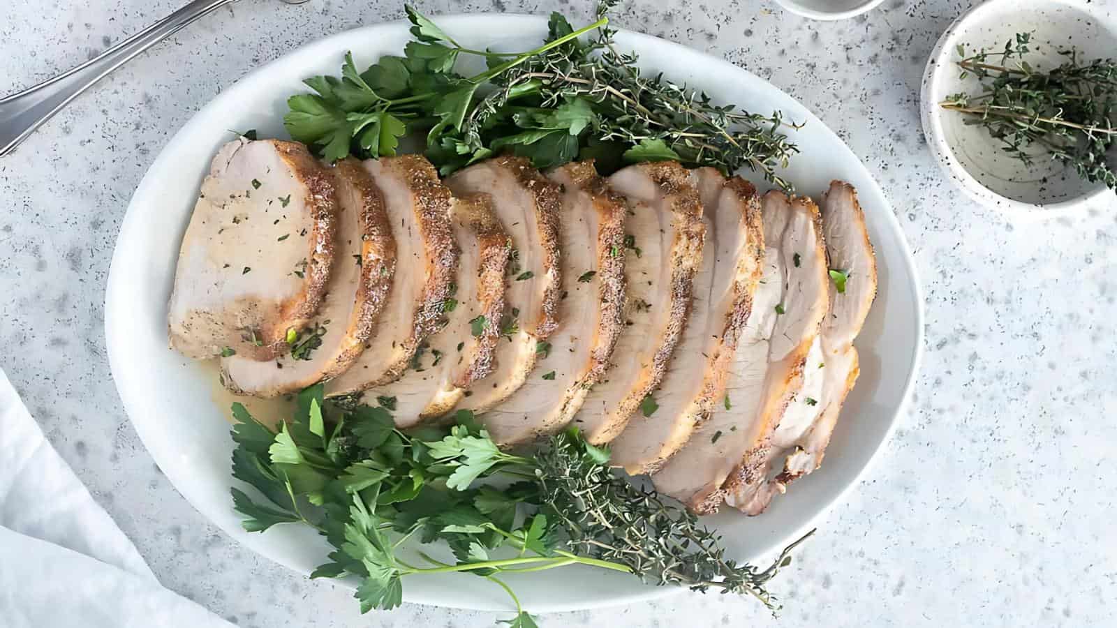 Sliced roasted pork loin garnished with fresh parsley and thyme on a white plate, placed on a speckled countertop.