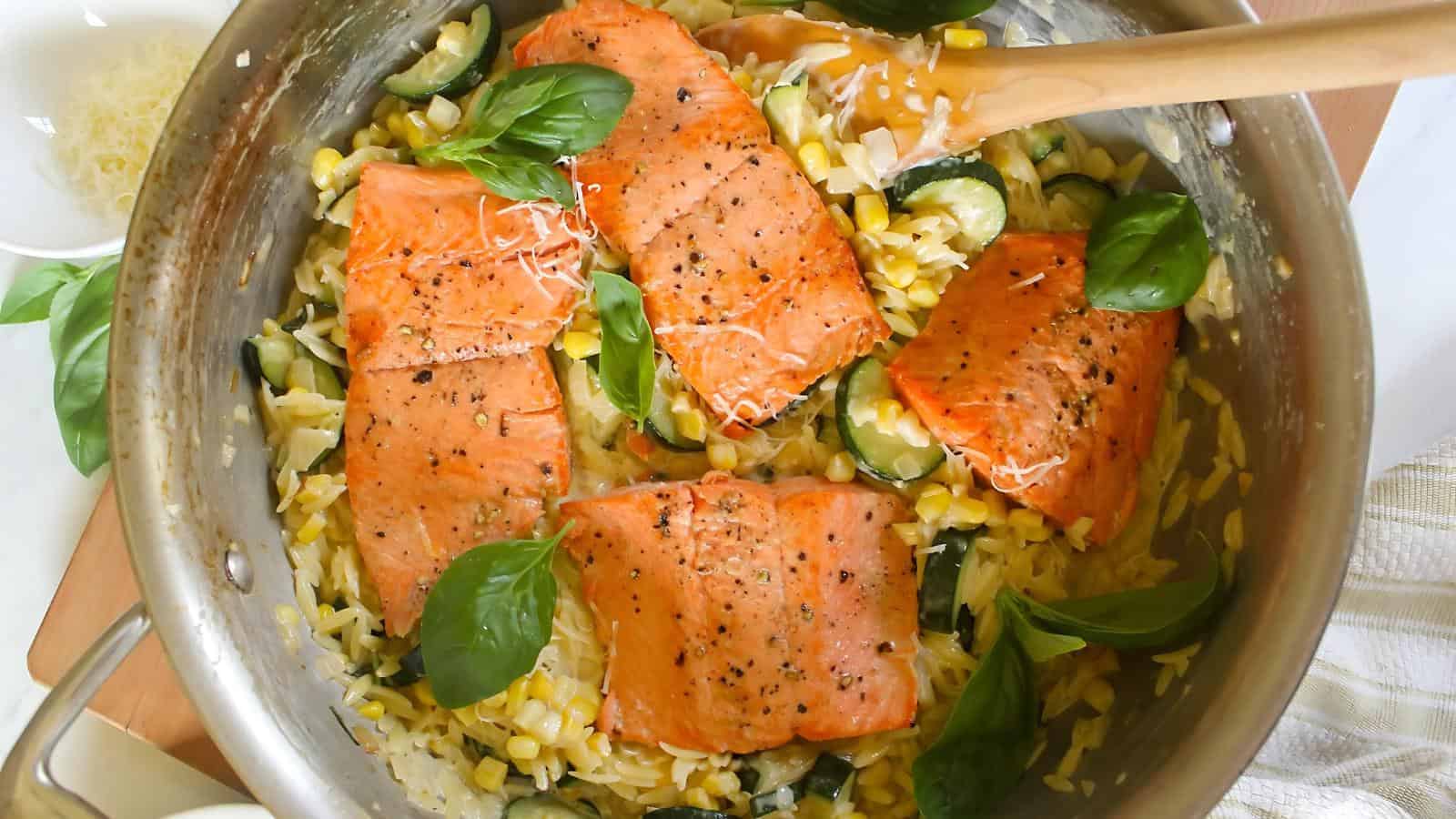 Salmon fillets with basil on top of orzo, corn, and zucchini in a pan, with a wooden spoon resting on the side.