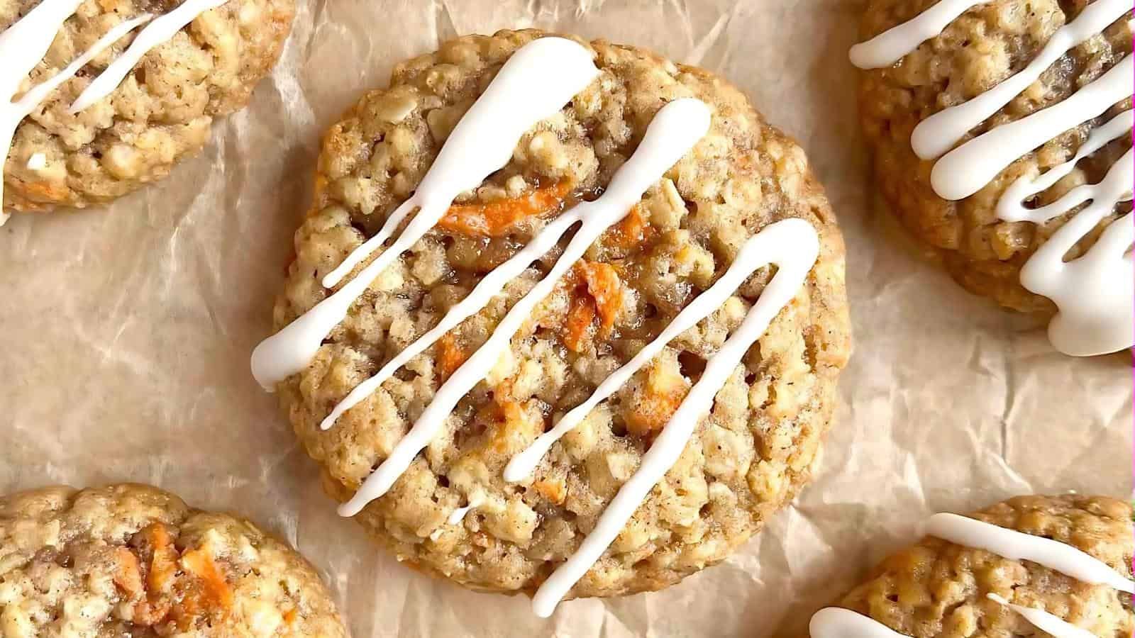 A close-up of oatmeal cookies with white icing drizzled on top, resting on brown parchment paper.
