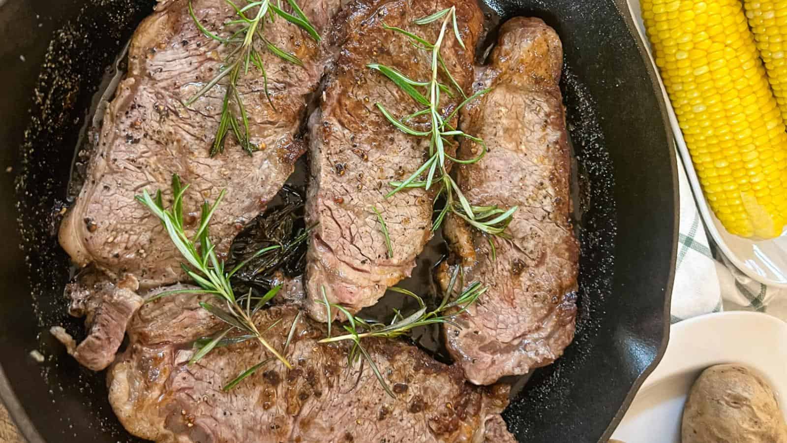 Four steaks with rosemary sprigs in a skillet. Corn on the cob and a potato are visible in the background.