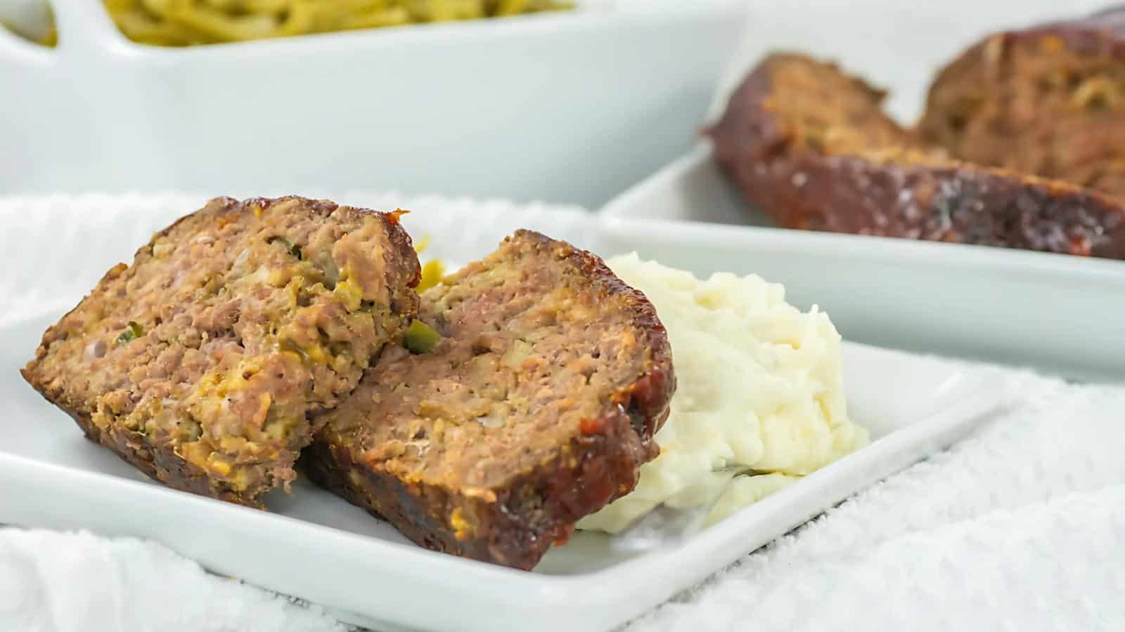 Slices of meatloaf served with mashed potatoes on a white plate, with a side of green beans in the background.