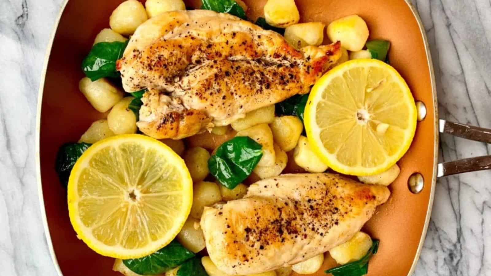 Pan with cooked chicken breasts, gnocchi, spinach, and two lemon slices on a marble surface.