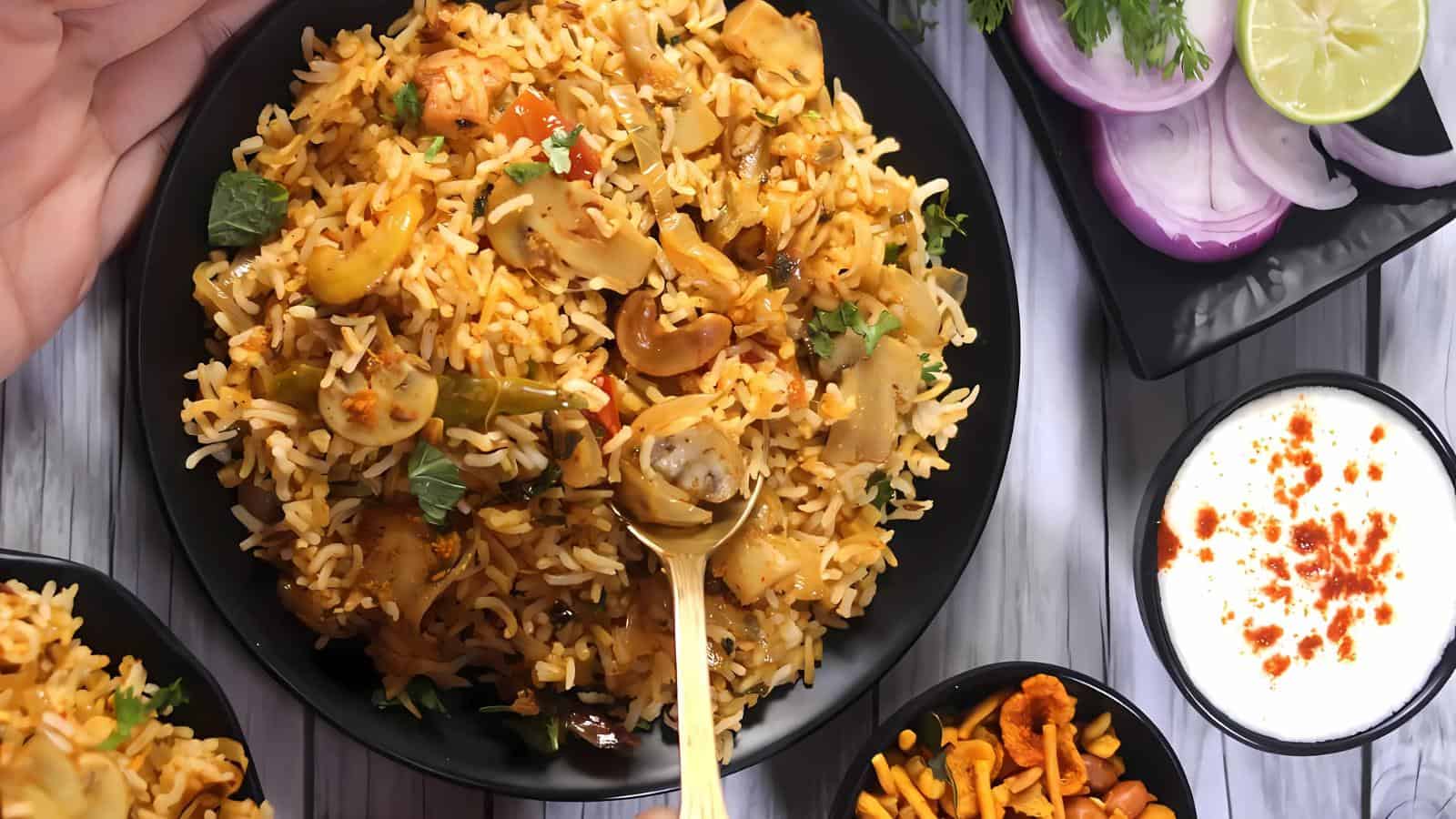 A bowl of biryani with vegetables and herbs is served with sliced onions, lime, yogurt, and a snack mix on the side.