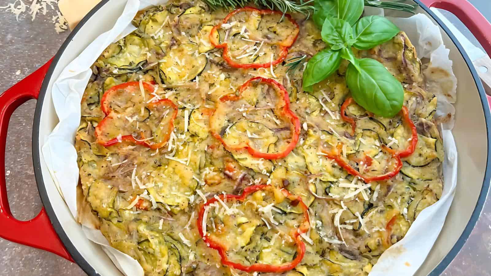 A baked vegetable dish with sliced red peppers, zucchini, grated cheese, and basil garnish on top.