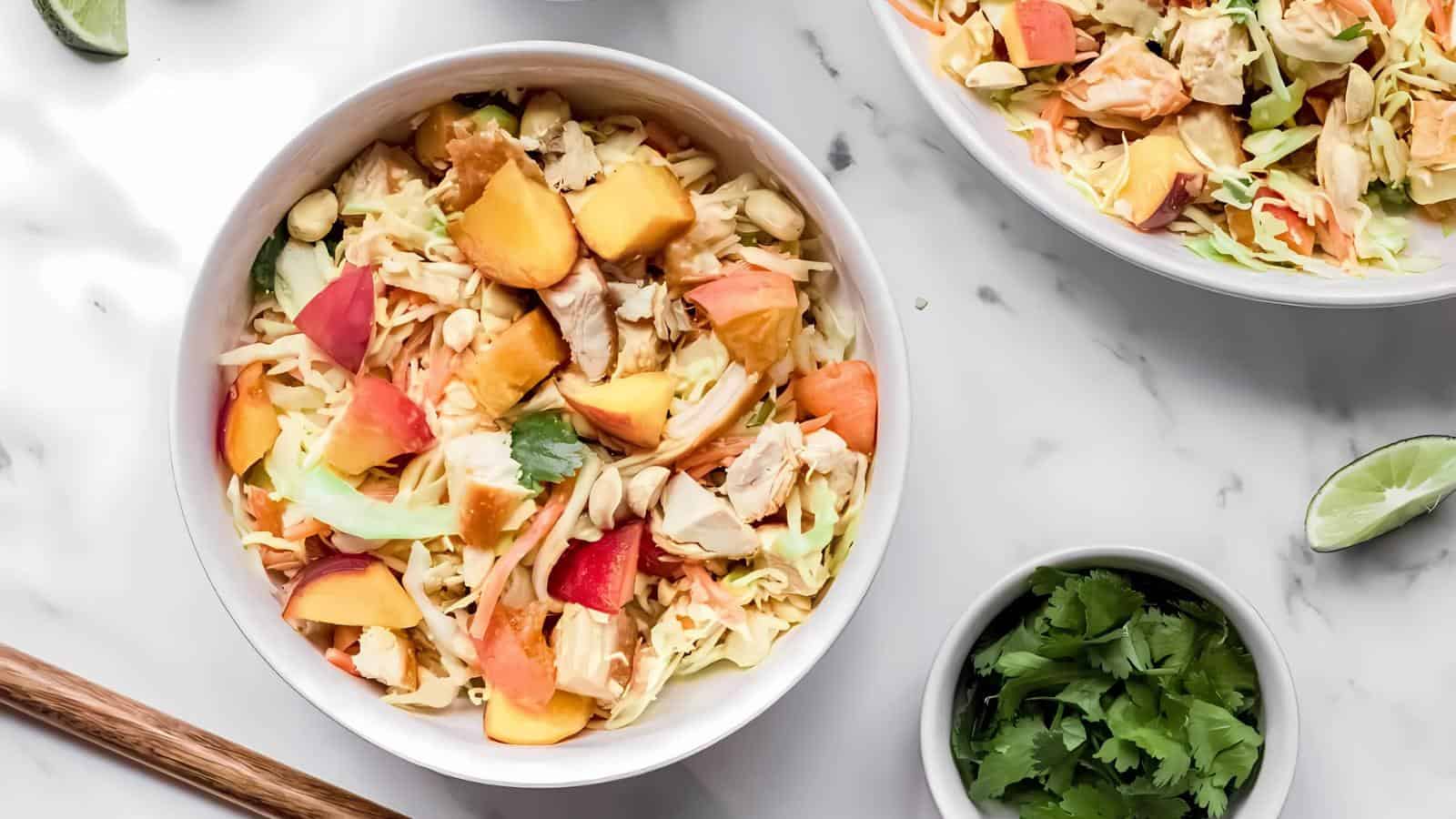 Bowl of salad with peach slices, shredded cabbage, and chicken pieces on a marble surface, accompanied by a bowl of cilantro and lime wedges.