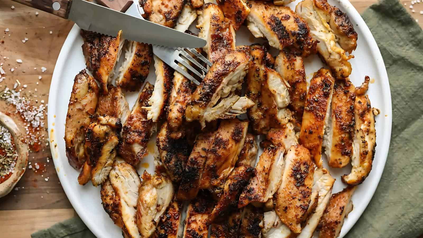 A plate of grilled chicken slices seasoned with spices. Knife and fork are placed on top. A green cloth is partially visible underneath.