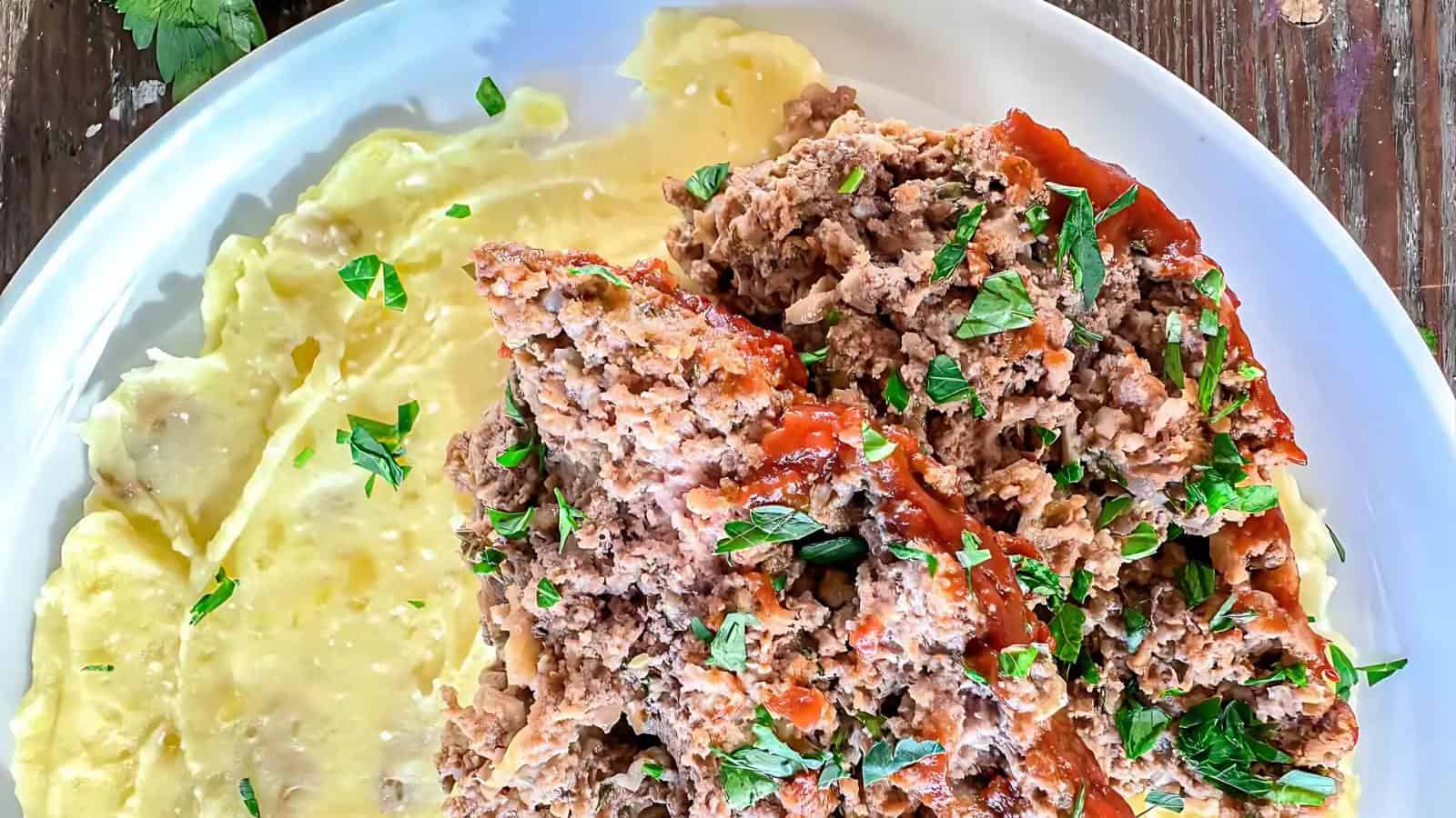 A plate with slices of meatloaf topped with herbs, next to a serving of creamy mashed potatoes with green garnish.